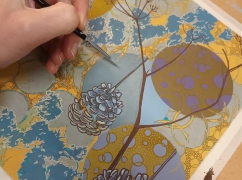 An image of a hand with a paintbrush working on an illustration of a pine cone, tree and blue/gold patterns.
