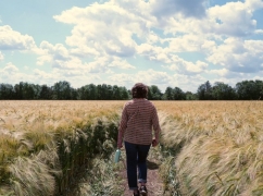 A person is in the centre of the foreground with their back to the camera, walking along a track through a large field, towards a horizon line of trees.
