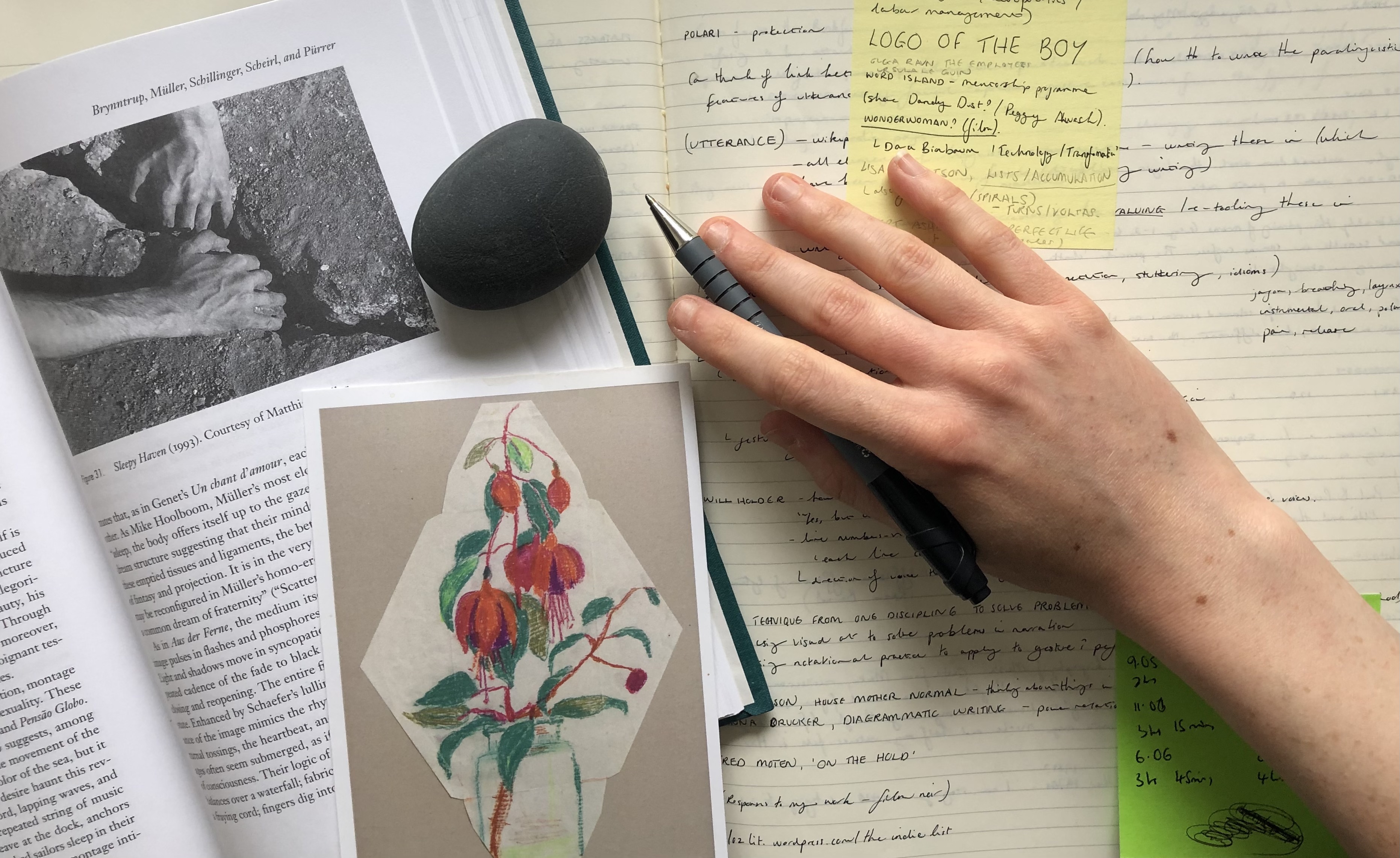 image shows a white person’s hand holding a pen over a notebook page covered in writing and post-it notes. Covering the left side of the page is a book, open on an image of hands prising apart a crack in a rock. Over the top of the book is a postcard of a drawing of flowers, and a small round grey pebble..